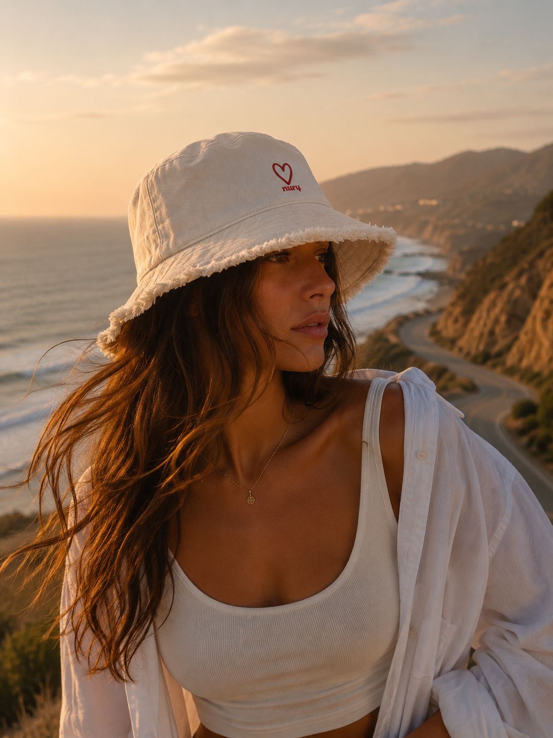 Model wearing a cream Nury bucket hat with the heart mark, coastal road at golden hour